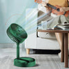 Green fan blowing air towards a person sitting on a couch with a glass of water and a book.